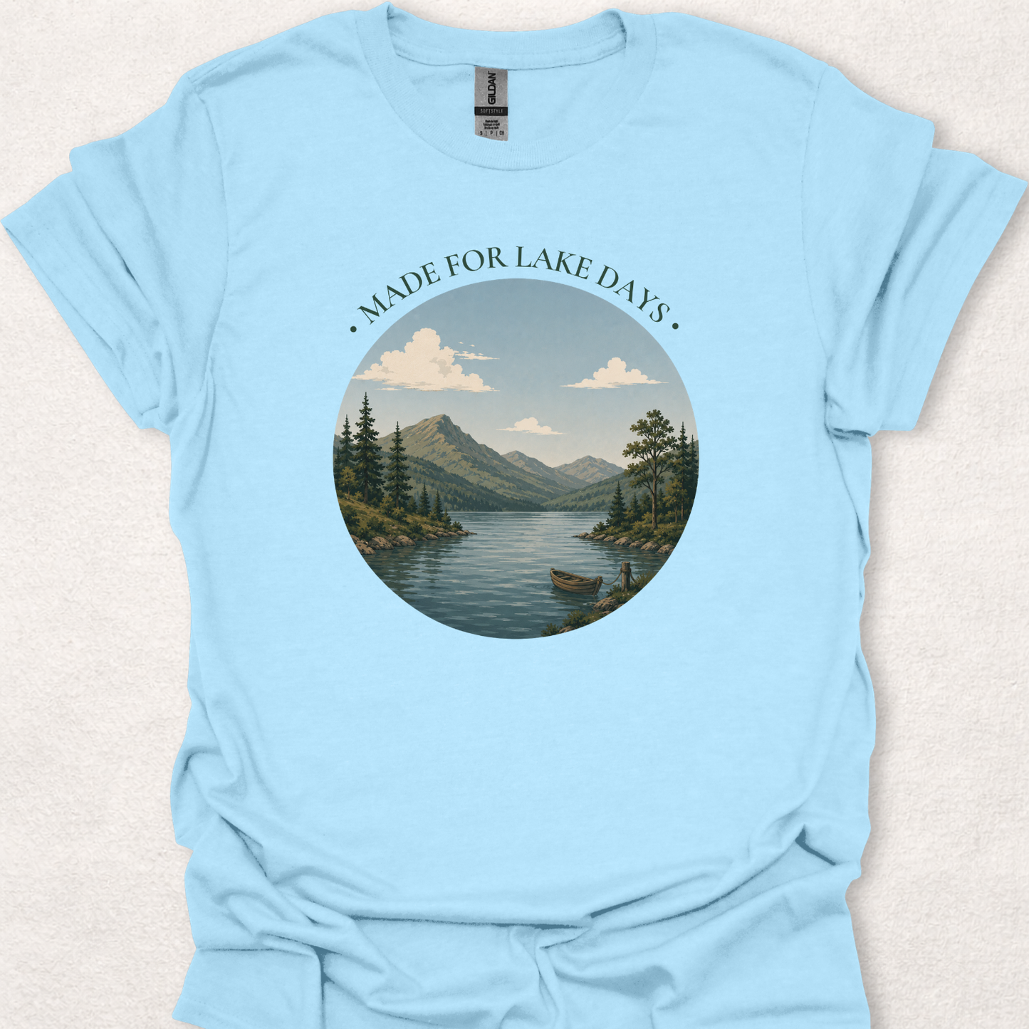 Light blue t-shirt featuring the Made For Lake Days design with a peaceful lakeside scene, mountain backdrop, evergreen trees, and a small boat.