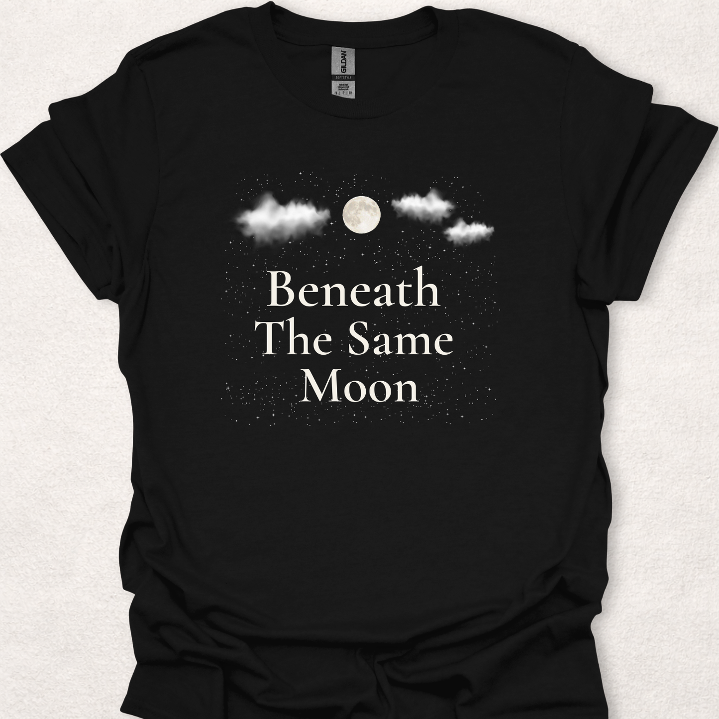 Nature-inspired moon and stars t-shirt featuring the phrase "Beneath The Same Moon" with a full moon, clouds, and a starry night sky.