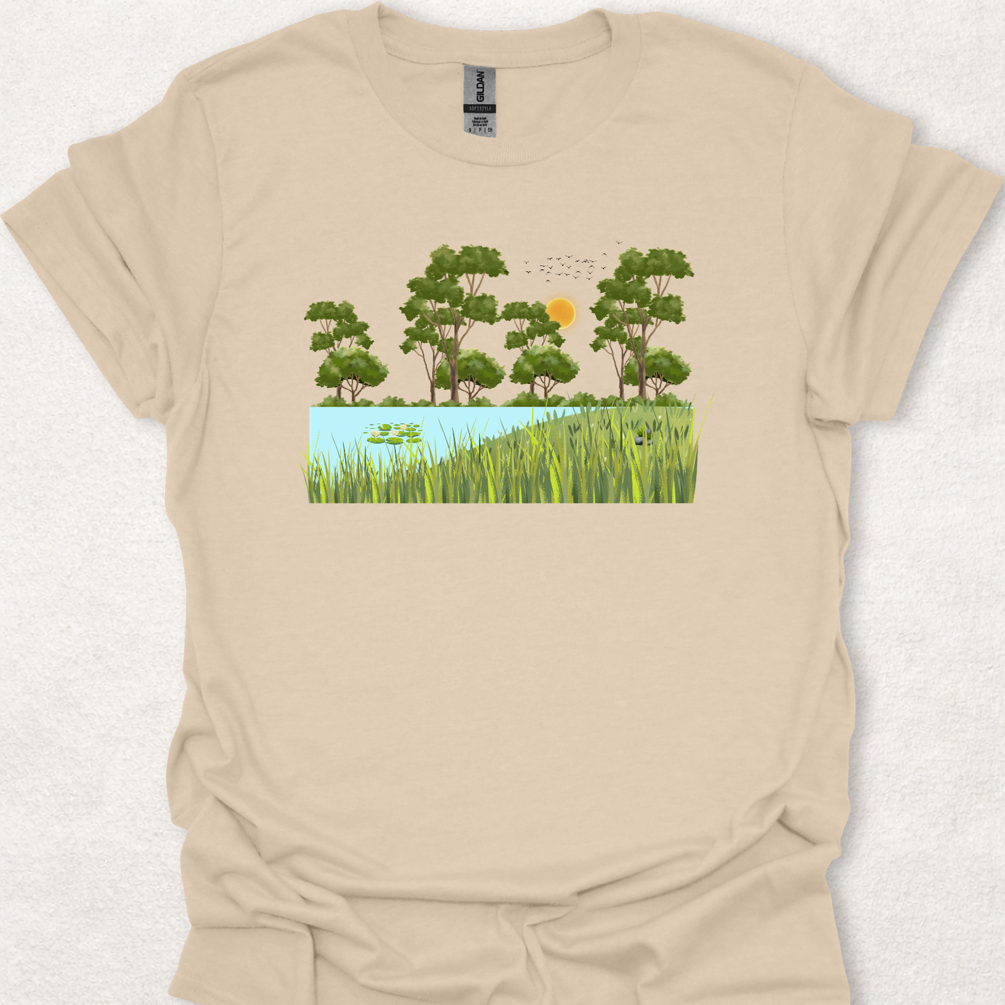 Peace Grows Here nature-inspired t-shirt in sand featuring a peaceful pond, trees, birds, and a sunny landscape scene.