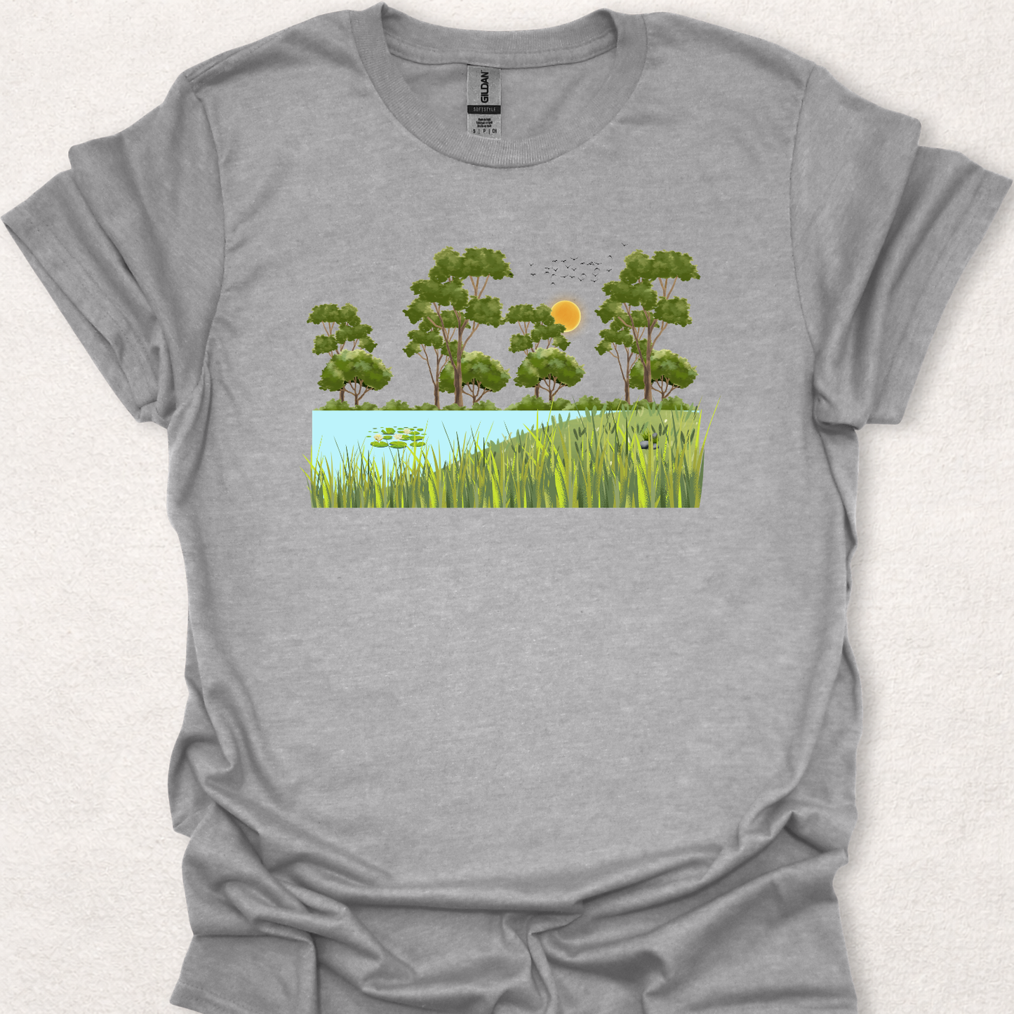 Peace Grows Here nature-inspired t-shirt in sand featuring a peaceful pond, trees, birds, and a sunny landscape scene.