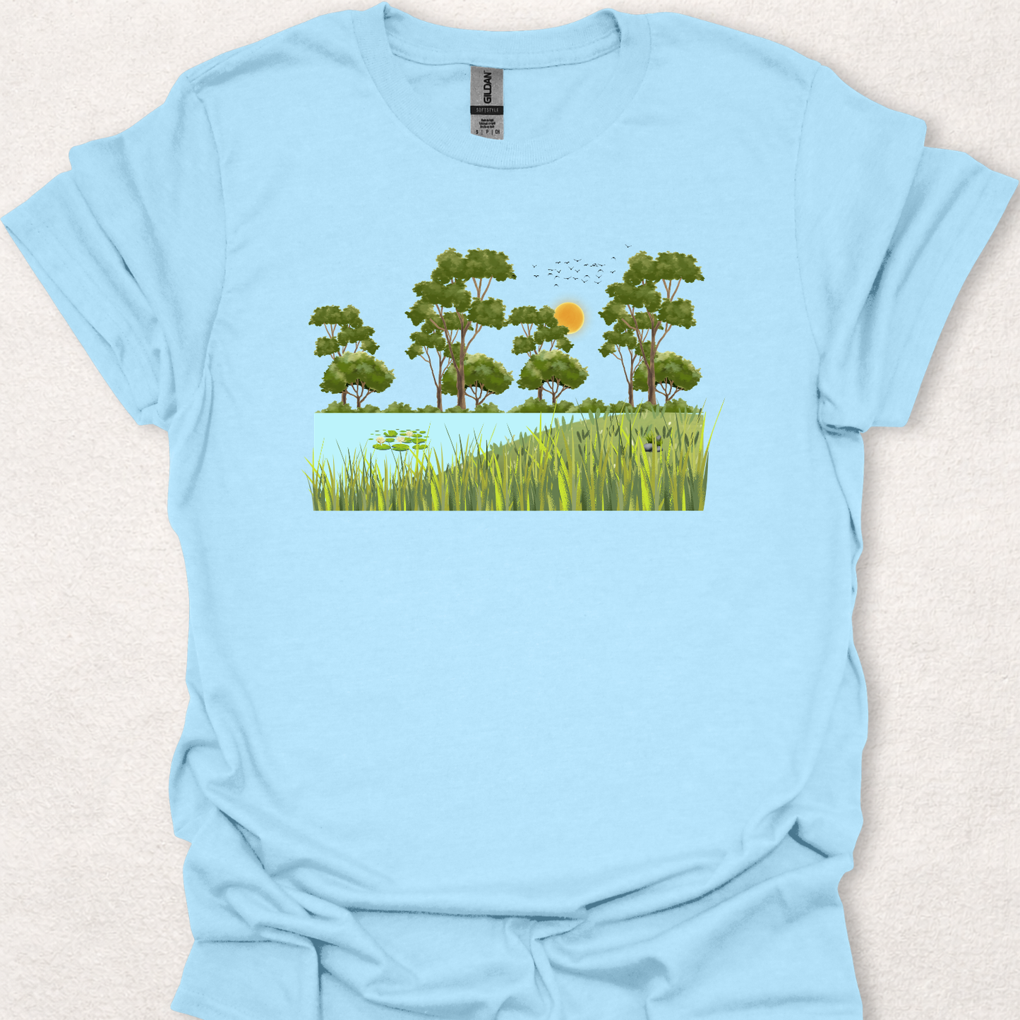 Peace Grows Here nature-inspired t-shirt in light blue featuring a peaceful pond, trees, birds, and a sunny landscape scene.