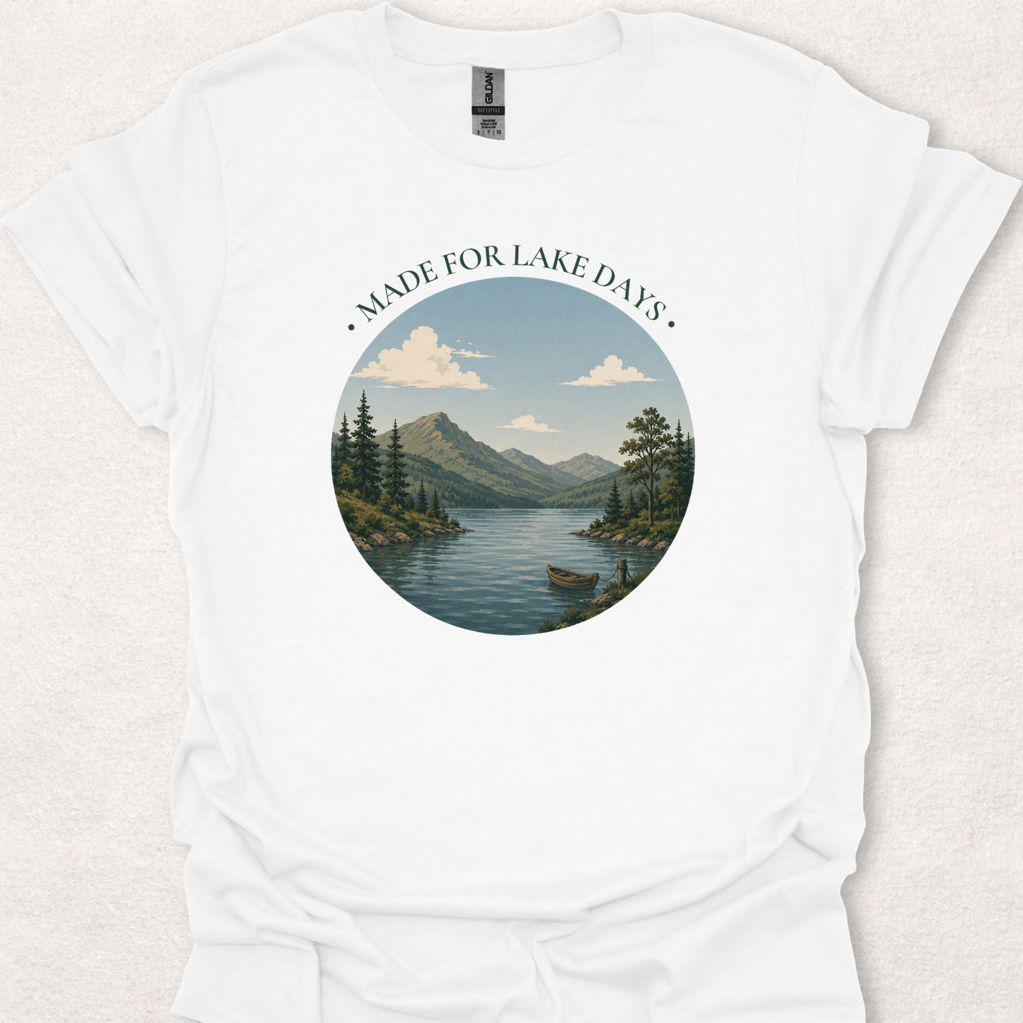 White t-shirt featuring the Made For Lake Days design with a serene lake landscape, mountains, evergreen trees, and a small boat.