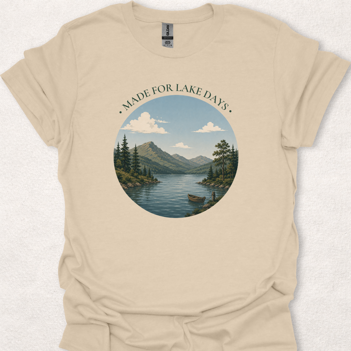 Sand-colored t-shirt featuring the Made For Lake Days design with a calm mountain lake scene, small boat, and evergreen trees.