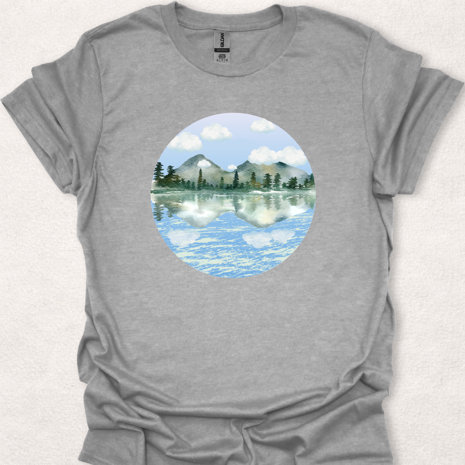 Stillwater nature-inspired t-shirt featuring a watercolor mountain lake with reflections of trees, mountains, and clouds.