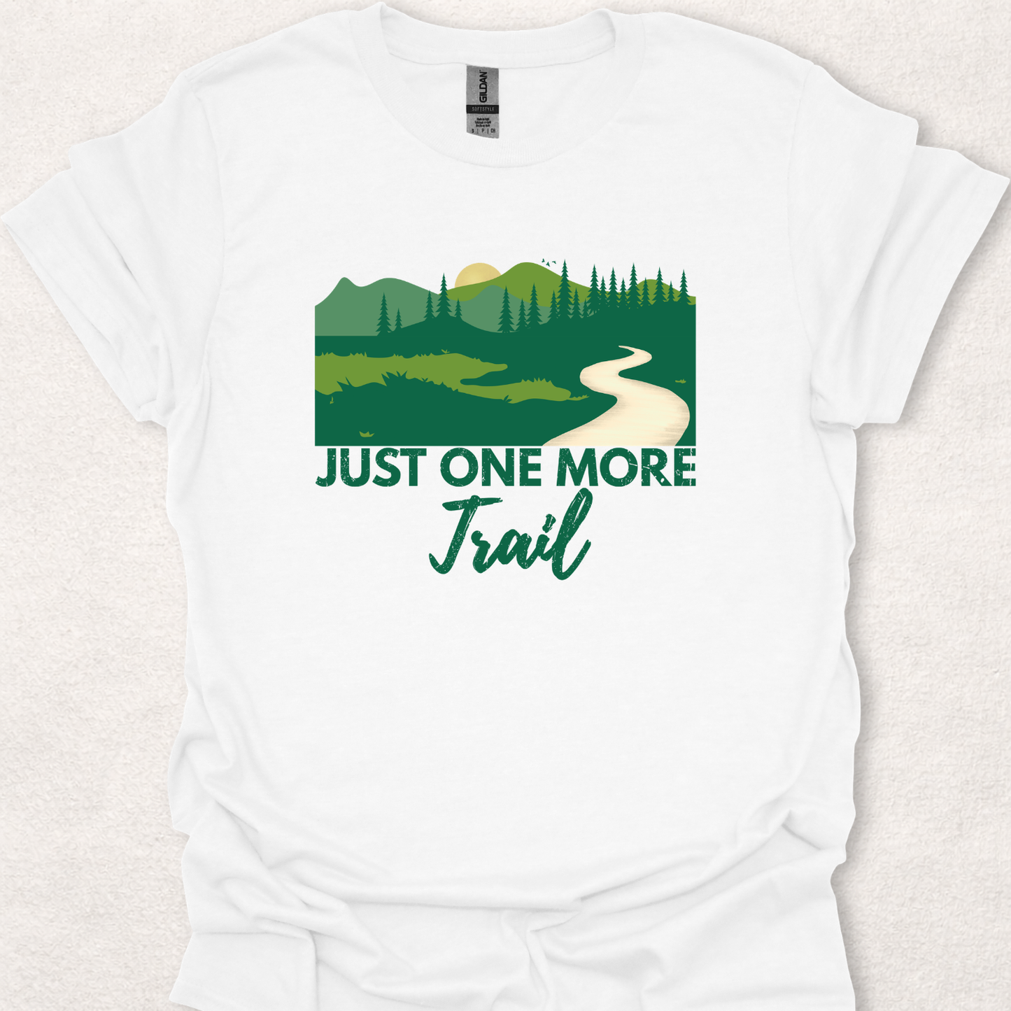 White unisex t-shirt featuring the "Just One More Trail" hiking design with a scenic forest landscape, mountains, trees, and winding trail for hikers and nature enthusiasts.