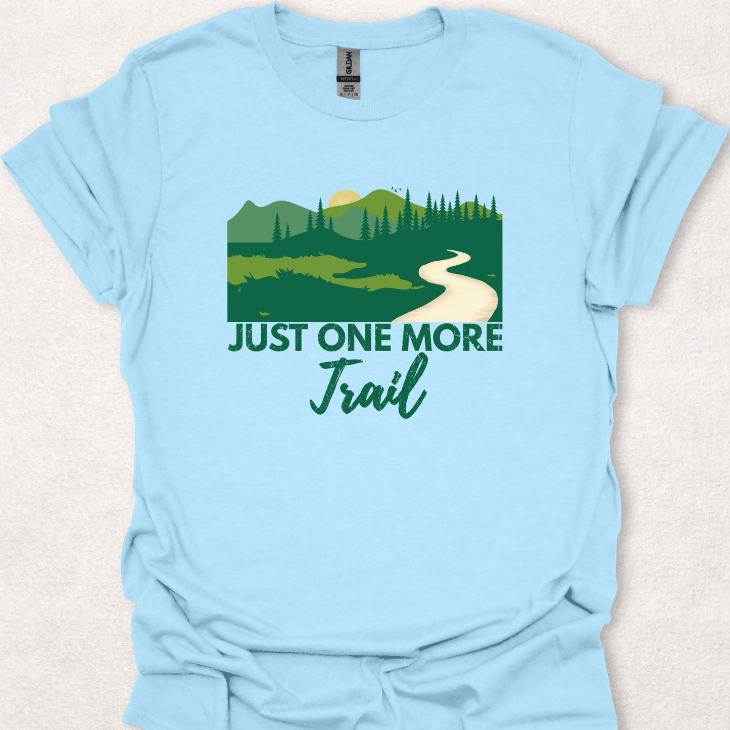 Light blue unisex t-shirt featuring the "Just One More Trail" hiking design with a scenic forest landscape, mountains, trees, and winding trail for nature lovers and outdoor adventurers.